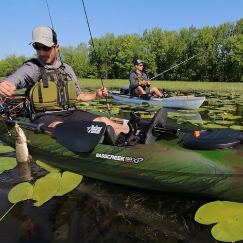 Two people kayak fishing on a lake with lily pads.