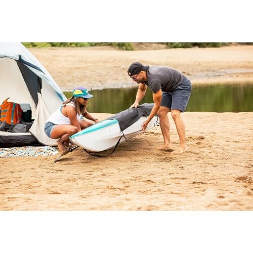 Two people setting up a kayak on a sandy beach near a tent.